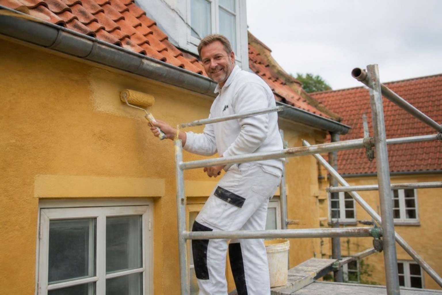 Smilende maler i hvidt arbejdstøj står på et stillads og ruller gul maling på en pudset facade med tegltag